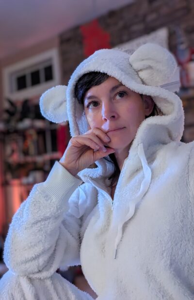 Woman in white onesie with mouse ears, hand on chin.